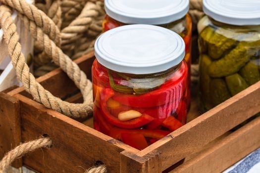 Wooden crate with glass jars with pickled red bell peppers and pickled cucumbers (pickles) isolated. Jars with variety of pickled vegetables. Preserved food concept in a rustic composition.