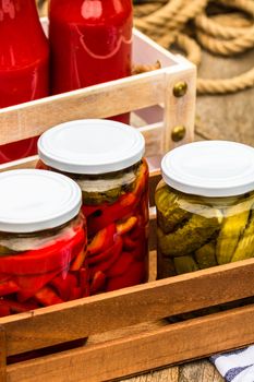 Wooden crate with glass jars with pickled red bell peppers and pickled cucumbers (pickles) isolated. Jars with variety of pickled vegetables. Preserved food concept in a rustic composition.