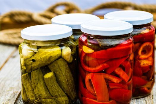 Glass jars with pickled red bell peppers and pickled cucumbers (pickles) isolated. Jars with variety of pickled vegetables. Preserved food concept in a rustic composition.