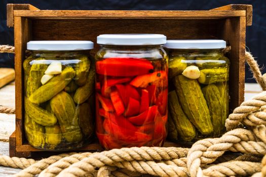 Glass jars with pickled red bell peppers and pickled cucumbers (pickles) isolated in wooden crate. Jars with variety of pickled vegetables. Preserved food concept in a rustic composition.