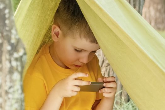 Boy playing video game outdoor in park, backyard, garden. Kid with phone in tent. 6 years old child having fun on nature. E-learning concept. Children and gadgets. Entertainment. Summer holidays