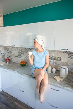 A girl with a towel on her head after a shower sits on the table in her kitchen and drinks coffee