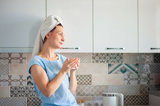 A woman washed her hair in the morning and drinks coffee with a towel on her head. Morning toilet and breakfast in the kitchen