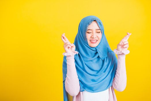 Asian Muslim Arab, Portrait of happy Asian beautiful young woman Islamic religious wear veil hijab she smiling and holding fingers crossed for good luck, studio shot isolated on yellow background