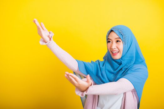 Asian Muslim Arab, Portrait of happy beautiful young woman Islam religious wear veil hijab funny smile she open arms for hug or raise hand to pick up items receive something from above isolated yellow