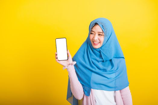 Asian Muslim Arab woman Islam wear veil hijab funny smile she talking on showing blank screen smart mobile phone, studio shot isolated on yellow background