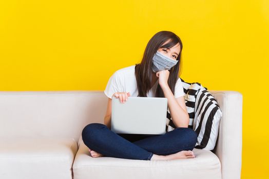 Portrait Asian of beautiful young woman sitting on sofa wearing medical face mask protective she work from home with laptop computer during Coronavirus studio shot isolated on yellow background