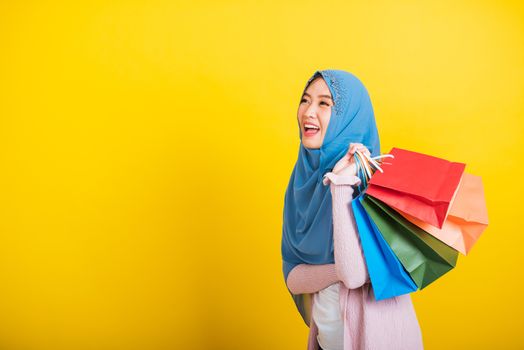Asian Muslim Arab, Portrait of happy beautiful young woman Islam religious wear veil hijab funny smile she holding colorful shopping bags, studio shot isolated on yellow background