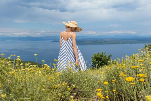 Rear view of young woman wearing striped summer dress and straw hat standing in super bloom of wildflowers, relaxing while enjoing beautiful view of Adriatic sea nature, Croatia.