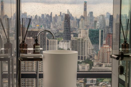 Modern white restroom with cityscape that can look outside from big window on a rainy day. Elegant faucets and components, Sanitary ware, The concept of health care. Selective focus.