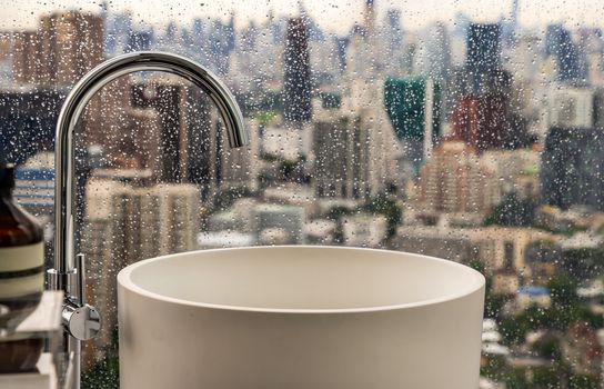 Modern hygienic hand basin with cityscape that can look outside from big window on a rainy day. Elegant faucets and components, Sanitary ware, The concept of health care. Selective focus.