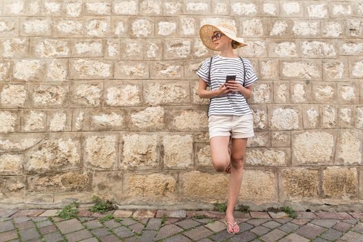 Beautiful young female tourist woman standing in front of old textured stone wall at old Mediterranean town, smiling, holding, smart phone to network on vacationes. Copy space.