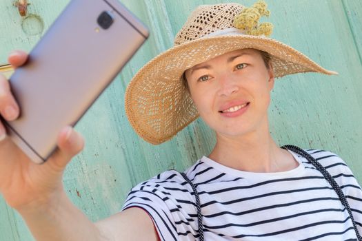 Beautiful young female tourist woman wearing big straw hat, taking self portrait selfie, standing in front of vinatage turquoise wooden door at old Mediterranean town.