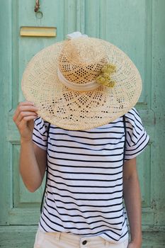 Unrecognizable female tourist woman wearing big straw standing in front of vinatage turquoise wooden door at old Mediterranean town.