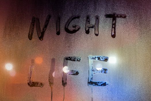 the words night life handwritten on night wet window glass surface - close-up with selective focus and background blur.