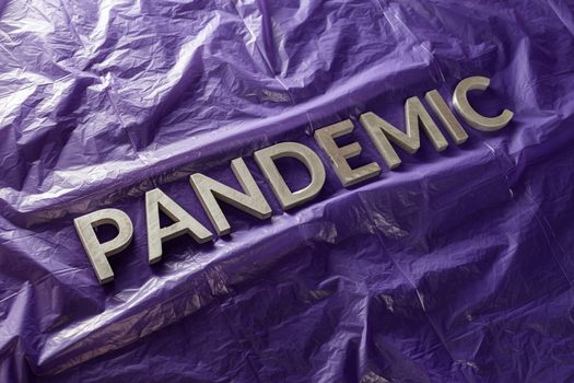 the word pandemic laid with silver metal letters on crumpled violet protective plastic film background in slanted diagonal perspective.