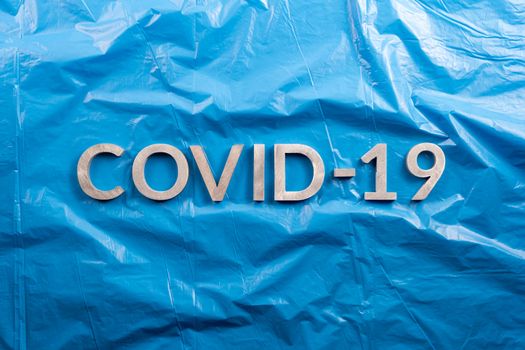 The word COVID-19 laid with aluminium letters on crumpled blue plastic film background in flat lay composition, directly above view.