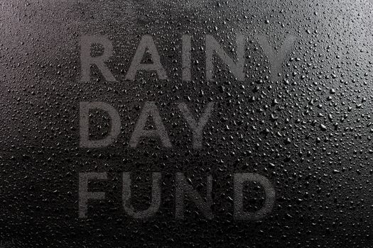 the dry words rainy day fund covered from water drops on black surface, unusual implementation concept in flat lay perspective.