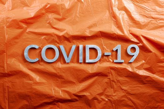 The word COVID-19 laid with aluminium letters on crumpled orange plastic film background in flat lay composition, directly above view.