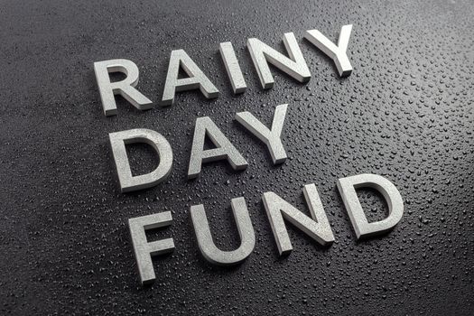 the words rainy day fund laid with silver metal letters on black background, all covered with water drops, slanted diagonal perspective with selective focus and blur