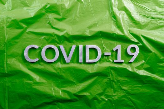 The word COVID-19 laid with aluminium letters on crumpled green plastic film background in flat lay composition, directly above view.