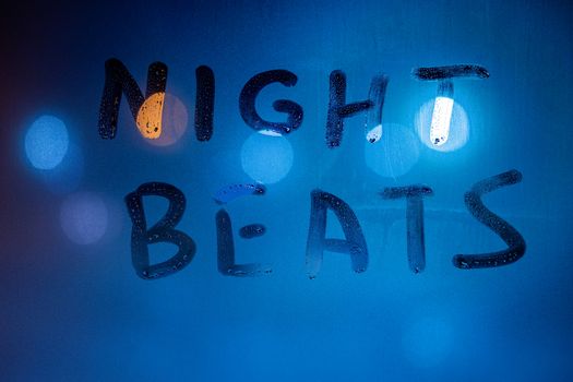 The words night beats handwritten on wet window glass at nighttime with blue light color. Close-up wuth shallow depth of field.