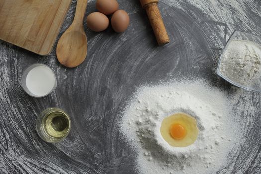 Broken chicken egg in a pile of flour, olive oil, milk, kitchen tool on gray table background. Products for baking bakery products. Cutting board, rolling pin, flour sieve, wooden spoon. bread or cake