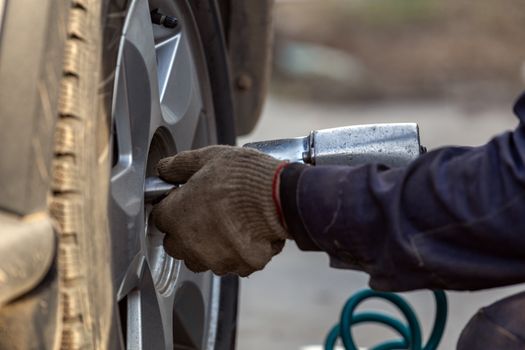 Hands of automotive mechanic unscrewing nuts with pneumatic impact wrench during car wheels season changing. Close-up view with selective focus and background blur.