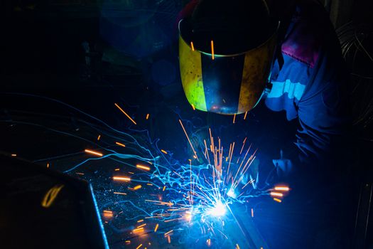 A process of manual operational semi-automatic electric welding with sparks, professional industrial worker in black-yellow protective mask at work.. High contrast dark close-up scene. MIG - metal inert gas - type of welding.