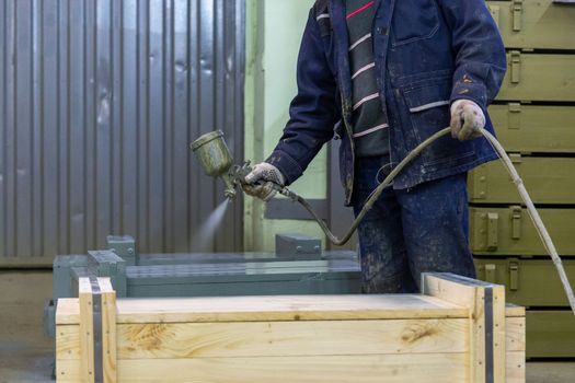 worker with airbrush painting big wooden crates to gray color.