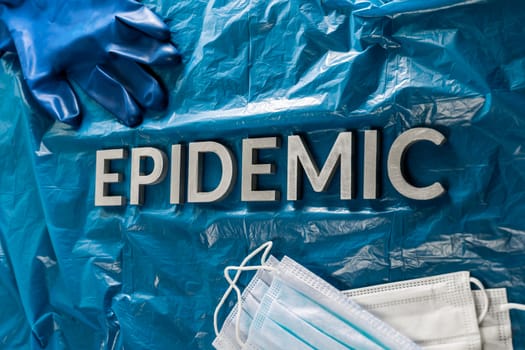 the word epidemic laid by metal letters in crumpled blue plastic background with face masks and blue protective glove.