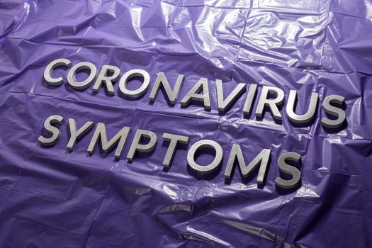 the words coronavirus symptoms laid with silver letters on crumpled purple plastic film - diagonal perspective composition with dramatic light