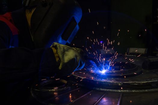 A lens flare while manual semi-automatic electric welding with sparks, professional industrial worker in black protective mask at work. High contrast dark close-up scene. MIG - metal inert gas - type of welding.