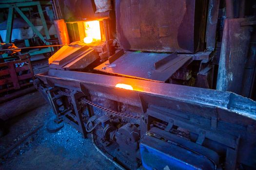 A process of moving luminous hot metal workpiece from furnace to hammer with old dirty industrial chain conveyor.