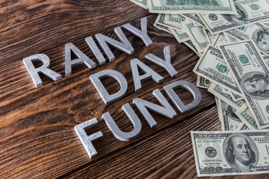 words RAINY DAY FUND laid on wooden surface with metal letters with rain drops and us dollar banknotes - diagonal perspective view