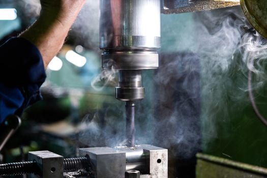 Industrial steel drilling with smoke close-up with selective focus. Manual feed by hand. Workpiece in vise on table. High contrast with background blur.