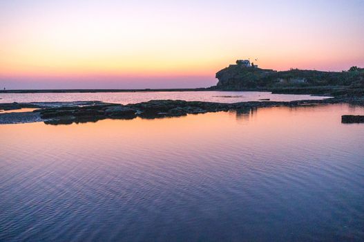 Sunset long exposure shot showing a calm sea lake lagoon with rocks in the foreground and a temple on an overlooking hill in the distance. Shows amazing locations in gujarat maharastra west coast of india