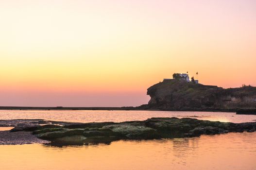 Sunset long exposure shot showing a calm sea lake lagoon with rocks in the foreground and a temple on an overlooking hill in the distance. Shows amazing locations in gujarat maharastra west coast of india