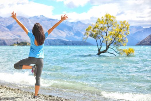 New Zealand morning yoga girl doing tree pose variation doing yoga practice with open arms at Wanaka lake by the lone tree, tourist travel popular attraction in New Zealand. Beach nature landscape.