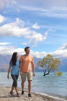 New Zealand tourists couple visiting Wanaka Lone Tree walking by lake. Happy young people traveling in summer adventure destination. Multiracial woman and man.