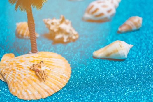 Miniature people wearing swimsuit relaxing on a seashell with blue glitter background