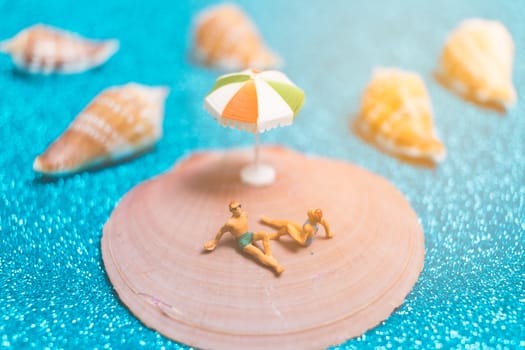 Miniature people wearing swimsuit relaxing on a seashell with blue glitter background