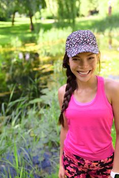 Happy smiling young Asian girl in sportswear sports cap and pink activewear portrait. Chinese Caucasian multiracial runner woman in her 20s in outdoor city park active lifestyle.