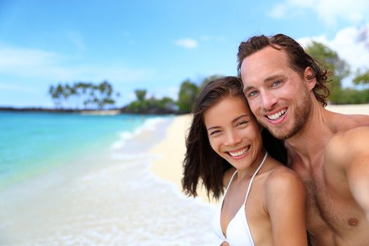 Happy beach selfie couple in love on Hawaii holidays. Healthy lifestyle young multiracial people smiling in swimwear on tropical vacation.