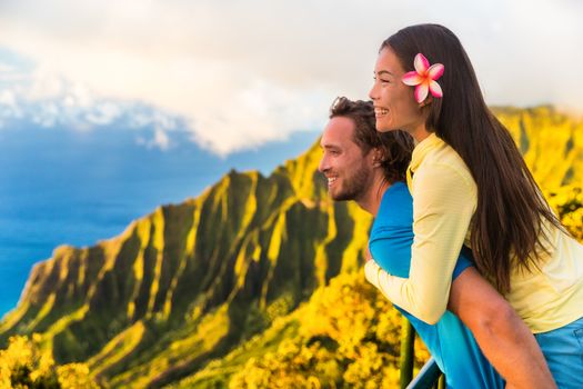 Travel couple fun piggyback at Na Pali Kauai Hawaii vacation adventure. Happy young Asian woman and Caucasian man piggybacking at nature lookout in mountains sunset. Love honeymoon vacations.