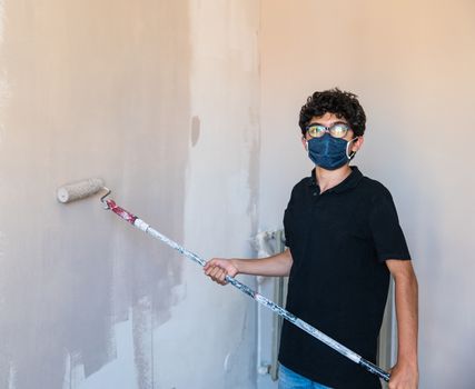 Renovate the walls of the house by painting them in the time of the coronavirus: the Caucasian boy has a mask to protect himself from the virus. Pass the impregnated roller of gray color on the wall.