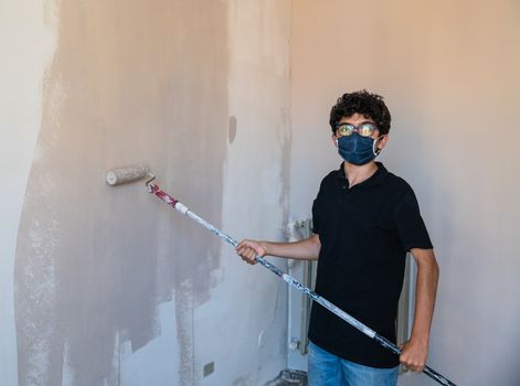 Renovate the walls of the house by painting them in the time of the coronavirus: the Caucasian boy has a mask to protect himself from the virus. Pass the impregnated roller of gray color on the wall.