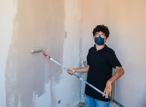 Renovate the walls of the house by painting them in the time of the coronavirus: the Caucasian boy has a mask to protect himself from the virus. Pass the impregnated roller of gray color on the wall.