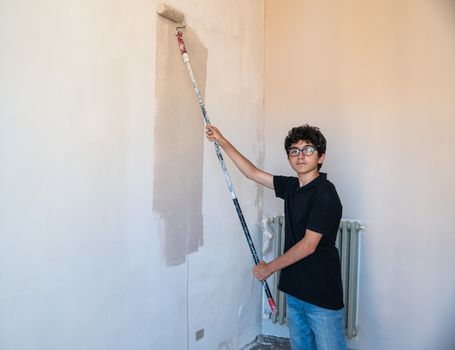 Renovate the walls of the house by painting them: the Caucasian boy with glasses passes the gray impregnated roller on the wall. A little color drips down the wall.