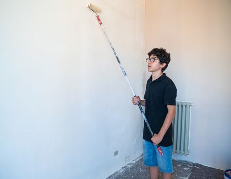 Renovate the walls of the house by painting them: the Caucasian boy with glasses passes the  roller on the wall